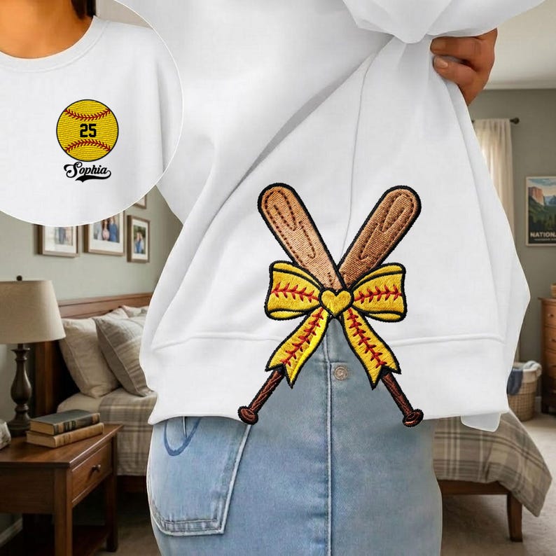 May include: White sweatshirt with embroidered softball and crossed bats design. The softball has the number 25 and the name "Sophia" below it. The crossed bats are brown with a yellow bow.