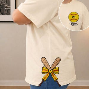 May include: Cream-colored t-shirt with a softball graphic and the name "Sophia." The shirt features crossed baseball bats with a yellow bow and heart design. The softball graphic includes the number 25.