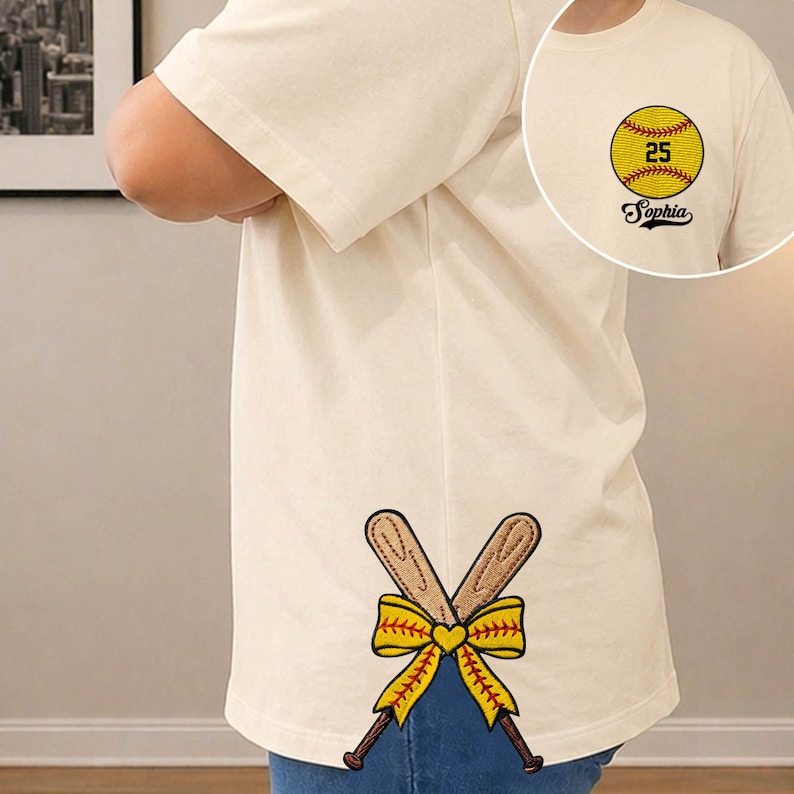 May include: Cream-colored t-shirt with a softball graphic and the name "Sophia." The shirt features crossed baseball bats with a yellow bow and heart design. The softball graphic includes the number 25.