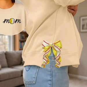 May include: Cream-colored sweatshirt with the word "mom" and a baseball graphic. A decorative yellow and white bow with red stitching is sewn onto the back of the sweatshirt. The sweatshirt is paired with blue jeans.
