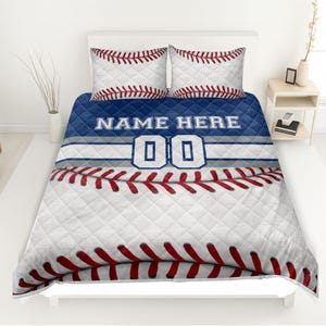 May include: A baseball-themed bedding set featuring a white and red baseball design. The set includes a quilt and two pillowcases. The quilt has a blue band with the text "NAME HERE" and the number "00".
