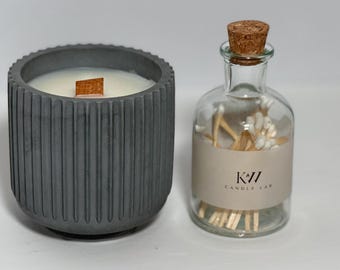 May include: A gray, ribbed concrete candle holder with a wooden wick next to a clear glass bottle with a cork stopper. The bottle contains matches and a label that reads "K77 CANDLE LAB".
