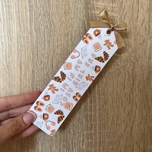 May include: A rectangular white bookmark featuring a repeating pattern of brown teddy bears, croissants, books, mugs, and slippers. The text "I'M IN MY COSY GIRL ERA" is printed in the centre. A gold satin ribbon is tied through a hole at the top.