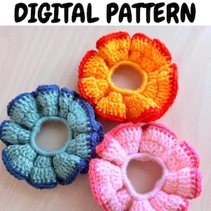 May include: Three handmade crochet scrunchies in shades of blue, orange, and pink, arranged on a light wood surface. The scrunchies are flower-shaped, with the text "CROCHET SCRUNCHIE DIGITAL PATTERN" at the top.