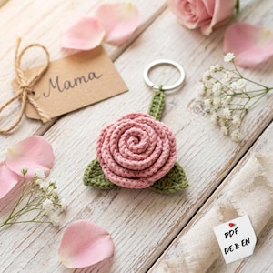 Könnte beinhalten: Ein handgefertigter, rosa gehäkelter Rosen-Schlüsselanhänger mit grünen Blättern. Der Schlüsselanhänger ist an einem silbernen Ring befestigt. Ein kleines Etikett mit dem Wort "Mama" ist mit Bindfaden gebunden. Rosa Rosenblätter und Blumen liegen verstreut.