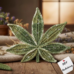 Peut inclure: Une feuille de cannabis crochetée à la main, dans des tons de vert et de blanc, ornée d'embellissements clairs en forme de gouttelettes. La feuille est posée sur une surface en bois, avec une note "PDF Anleitung".