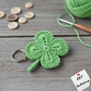May include: A handmade green crochet four-leaf clover keychain. The clover is made of yarn and attached to a silver-colored metal ring. A ball of green yarn, a crochet hook, and wooden buttons are in the background.