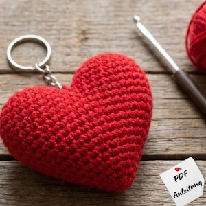 Puede incluir: Un llavero de corazón rojo tejido a mano con un anillo plateado. Un ganchillo y una madeja de hilo rojo están en el fondo. El llavero está sobre una superficie de madera. Un pequeño papel blanco con el texto "PDF Anleitung" también está en la imagen.