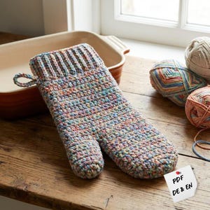 May include: A handmade, multi-colored crochet oven mitt with a loop for hanging. The mitt is made of yarn in shades of blue, pink, yellow, and green. A brown baking dish and balls of yarn are in the background. A small white tag with the text "PDF DE & EN" is attached.