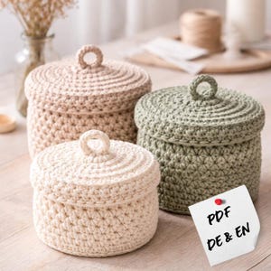 Puede incluir: Tres cajas de almacenamiento de ganchillo hechas a mano en crema, beige y verde salvia. Cada caja tiene una tapa a juego con un asa circular. Las cajas se exhiben sobre una superficie de color claro. Una nota con el texto "PDF DE & EN" está fijada.