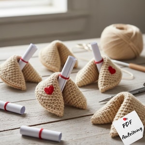 Può includere: Biscotti della fortuna all'uncinetto beige con cuori rossi. Ogni biscotto contiene una pergamena legata con filo rosso. Un gomitolo di lana, forbici e un uncinetto suggeriscono un progetto fai-da-te.