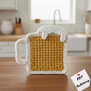 May include: A crocheted beer mug coaster with a yellow center, white foam detail, and a white handle. The coaster is square-shaped and sits on a wooden surface. A small note with the text "PDF Anleitung" is pinned to the surface.