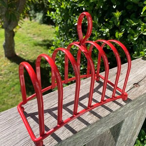 Puede incluir: Un tostador de metal rojo brillante con un asa. El soporte tiene seis ranuras curvas para sostener la tostada. Está colocado sobre una superficie de madera con un fondo verde.