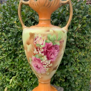 Könnte beinhalten: Eine antike Vase mit breitem Sockel und zwei Henkeln. Die Vase ist mit einem Blumendesign mit rosa und roten Rosen bemalt. Die Vase hat einen pfirsichfarbenen Sockel und eine Oberseite mit goldenen Akzenten an den Henkeln.