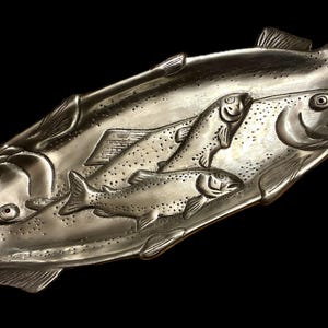 Puede incluir: Una fuente plateada con forma de pez. La fuente presenta un relieve detallado de tres peces, con dos peces más grandes formando la base y un pez más pequeño en medio. Sobre un fondo negro.