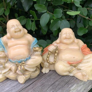 May include: Two smiling Buddha statues. The statues are cream-colored with gold accents. One Buddha wears a blue and gold robe and holds a gold object. The other Buddha wears an orange robe and holds a gold beaded necklace. The statues are sitting on a wooden surface.