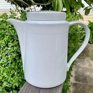 Op de afbeelding: Een witte theepot van keramiek met een eenvoudig ontwerp en een gladde, glanzende afwerking. De theepot heeft een rond lichaam en een tuit aan de ene kant. De handgreep is bevestigd aan de bovenkant van de theepot en buigt naar de andere kant.