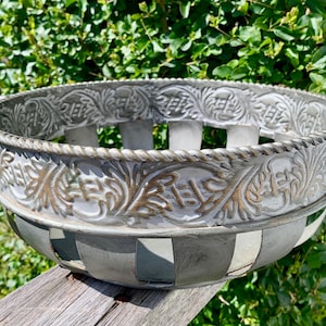 May include: A decorative metal planter with a distressed finish. The planter features a detailed floral pattern around the rim and a woven rope-like handle. The body has a gray and white checkered design.