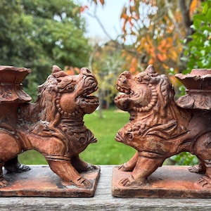 May include: Two terracotta Foo Dog candle holders. Each dog is in a crouching position with an open mouth, and a decorative candle cup on its back. The background is a blurred view of green foliage.