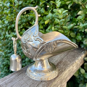 May include: Silver sugar bowl with a handle and a small bell. The bowl has floral and leaf patterns, a curved rim, and a pedestal base. The handle is arched, and the bell hangs from a chain. The bowl is on a wooden surface.