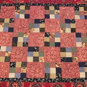 Puede incluir: Un edredón de retazos con un estampado floral en tonos de rosa, azul y crema. El edredón presenta un diseño de bloques repetidos con un borde floral negro. El edredón se muestra sobre una superficie con estampado de cachemira roja.