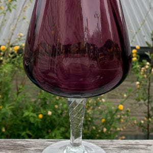 May include: A large wine glass with a deep purple bowl and a clear, twisted stem. The glass has a wide, ruffled rim and sits on a clear, circular base. The glass is set on a wooden surface with a blurred green background.