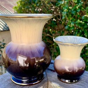 Puede incluir: Dos jarrones de cerámica con un diseño degradado. El jarrón más grande es de color morado oscuro a crema con un borde dorado. El jarrón más pequeño es de diseño similar pero de menor tamaño.