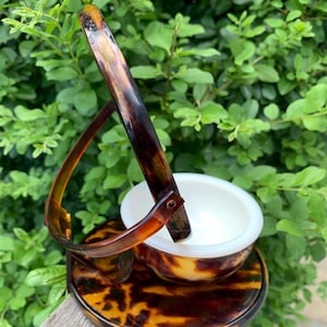 May include: A tortoiseshell  makeup holder with a hinged lid and a small white bowl inside. The holder has a tortoiseshell handle.