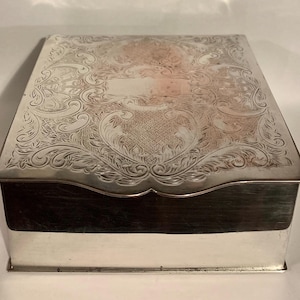 May include: A rectangular silver box with an engraved lid. The lid is decorated with detailed floral and scroll designs, and a central blank cartouche. The box has a dark base, possibly wood, and a polished silver finish.