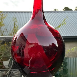 May include: A large, round, red glass vase with a narrow neck. The vase reflects the surrounding environment, including a house and trees. The vase is sitting on a wooden surface.