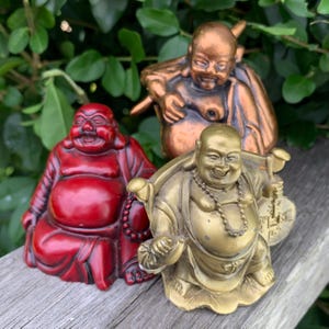 May include: Three smiling Buddha statues in red, bronze, and gold. The red Buddha is seated, the bronze Buddha is leaning, and the gold Buddha is holding a coin. The statues are on a wooden surface with green foliage in the background.