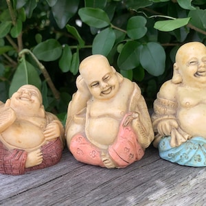 May include: Three smiling Buddha statues in various poses. The statues are beige with colorful accents of red, pink, and blue. The statues are sitting on a wooden surface with green foliage in the background.