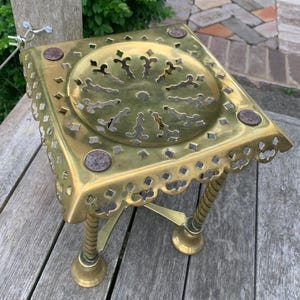 May include: A vintage brass table with a square top featuring decorative cut-out patterns. The table has three legs with a spiral design and decorative feet. The top has a circular design with cut-out shapes and four circular accents.