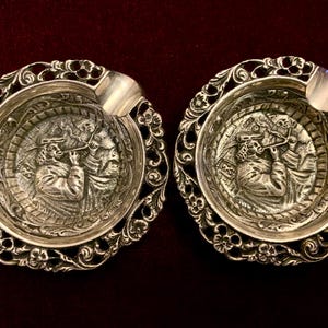 May include: A pair of silver ashtrays with a decorative floral edge and a central relief showing a person playing a wind instrument. Each ashtray has a small rest for cigarettes, set against a dark red background.