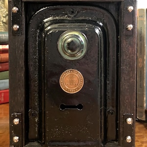 May include: A vintage-style, black wooden box with a central, glossy black panel. The panel features a circular, green-tinted glass element and a copper coin. The box is framed with dark wood and metal accents.