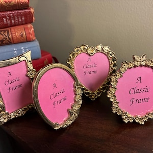 May include: Four gold-toned metal picture frames with ornate floral and butterfly detailing. Each frame contains a pink insert with the black text A Classic Frame. The set includes one rectangular, one oval, one heart-shaped, and one round frame.
