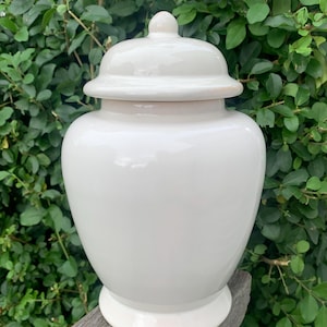 May include: A white ceramic ginger jar with a lid. The jar has a rounded body and a pedestal base. The lid is domed with a small knob on top. The jar is sitting on a wooden surface with green foliage in the background.