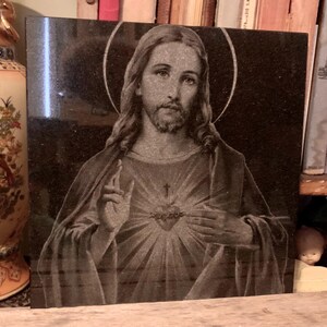 May include: A black granite plaque featuring a detailed engraving of Jesus Christ. The engraving depicts Jesus with his hands clasped over his heart, which is surrounded by a crown of thorns and a cross. The background is a dark, textured surface.