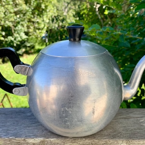 May include: A vintage aluminium teapot with a round body, a curved spout, and a black handle and lid knob. The teapot sits on a wooden surface with a green, leafy background.