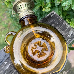 May include: An amber glass oil lamp with a brass burner and handle. The round body has a starburst design on the base. The lamp sits on a weathered wooden surface, with green foliage in the background.