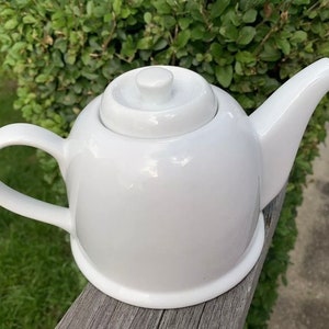 May include: A white ceramic teapot with a rounded body and a spout. The teapot has a lid with a knob on top.