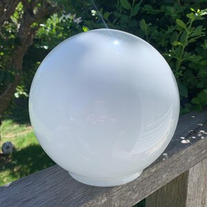 Puede incluir: Una gran esfera blanca opaca, posiblemente una lámpara, descansa sobre una superficie de madera. La esfera es lisa y reflectante, con una pequeña base. El fondo presenta follaje verde y luz solar.