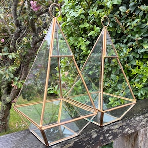 May include: Two geometric terrariums with clear glass panels and thin copper-toned metal frames. Each container features a pyramid shape with a square base and a small circular ring at the top for hanging or carrying purposes.
