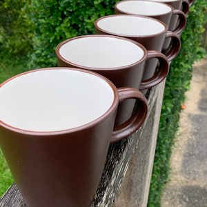 May include: A row of brown ceramic mugs with white interiors. The mugs are lined up on a wooden surface.