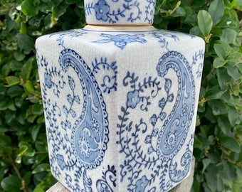 Peut inclure: Un pot en céramique carré avec un couvercle, avec un fond blanc et un motif cachemire bleu. Le pot est décoré de motifs floraux et repose sur une surface en bois. Le pot mesure environ 15 cm de haut.