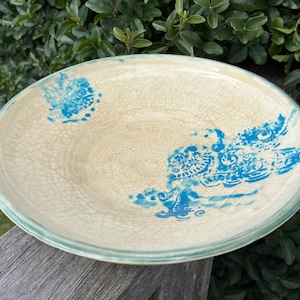 May include: A shallow ceramic bowl with a cream-colored base featuring a fine crackle glaze finish. The interior surface is decorated with vibrant blue abstract floral and scroll patterns applied in a stamped or painted style.