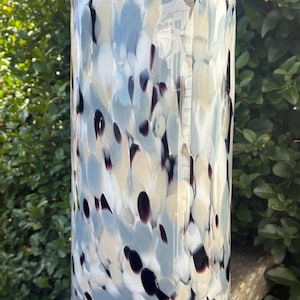 May include: A tall, cylindrical glass vase with a speckled design. The vase has a clear base and is decorated with blue, cream, and dark purple spots. The vase is set against a backdrop of green foliage.