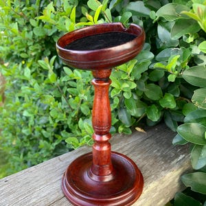 May include: A dark reddish-brown wooden pipe stand with a round base and a pedestal. The top has a circular indentation lined with black felt. The stand is displayed outdoors against a backdrop of green foliage.