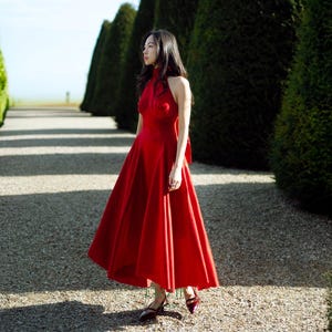 May include: A woman in a striking red halterneck dress with a full skirt, walking on a gravel path. The dress has a bow at the back. The setting is an outdoor space, with trimmed hedges and trees bordering the path.