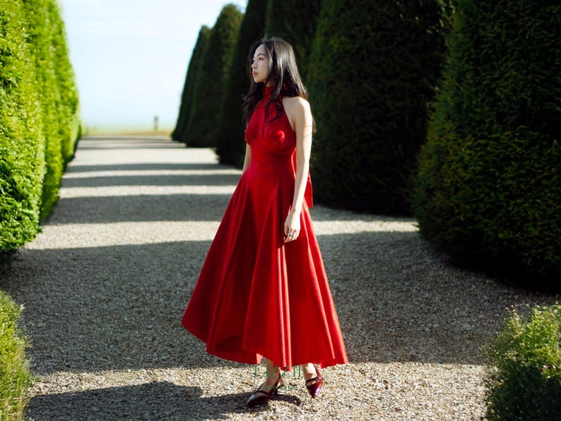 May include: A woman in a striking red halterneck dress with a full skirt, walking on a gravel path. The dress has a bow at the back. The setting is an outdoor space, with trimmed hedges and trees bordering the path.